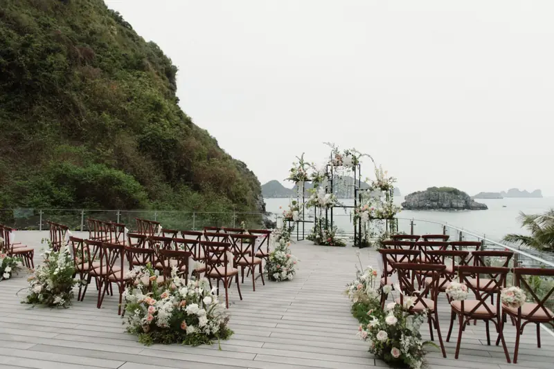 Sàn tiệc ngoài trời bày ghế gỗ và hoa, nhìn ra biển và đảo đá Cát Bà