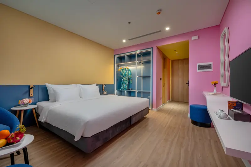 Phòng Deluxe Flamingo Hải Tiến: giường King, tường hồng – vàng, góc ngồi ghế đôn xanh