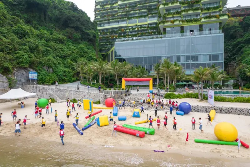 Đoàn khách chơi trò vận động với dụng cụ hơi nhiều màu trên bãi Cát Cò, phía sau là tòa khách sạn phủ cây xanh của Flamingo Cát Bà