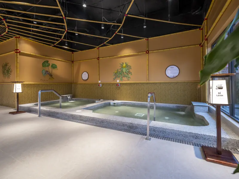 Hai bể onsen trong nhà có tay vịn inox, tường vàng ấm, trần tre đan