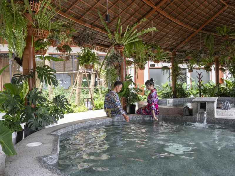 Hai khách mặc áo choàng thổ cẩm ngồi ngâm chân bên bể onsen ngoài trời dưới mái lá