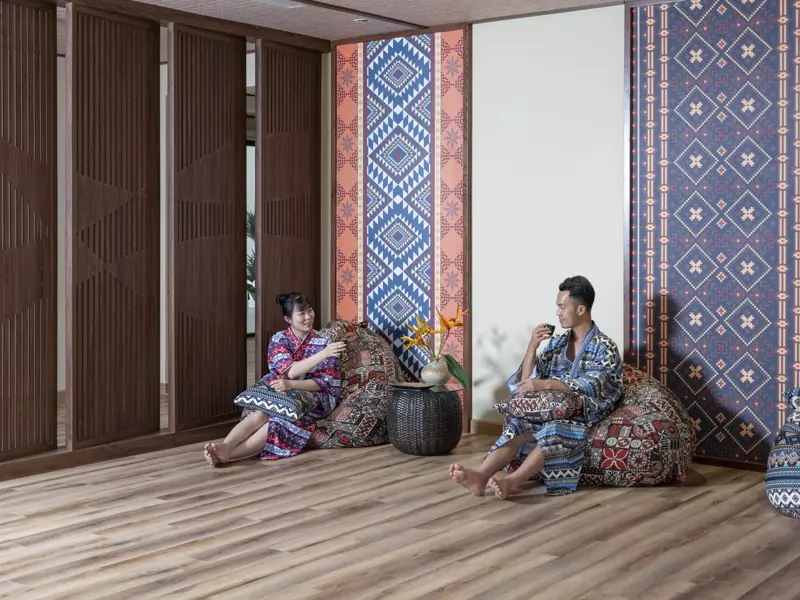 Hai khách ngồi nghỉ trên gối thổ cẩm trong khu thư giãn, vách hoa văn dân tộc