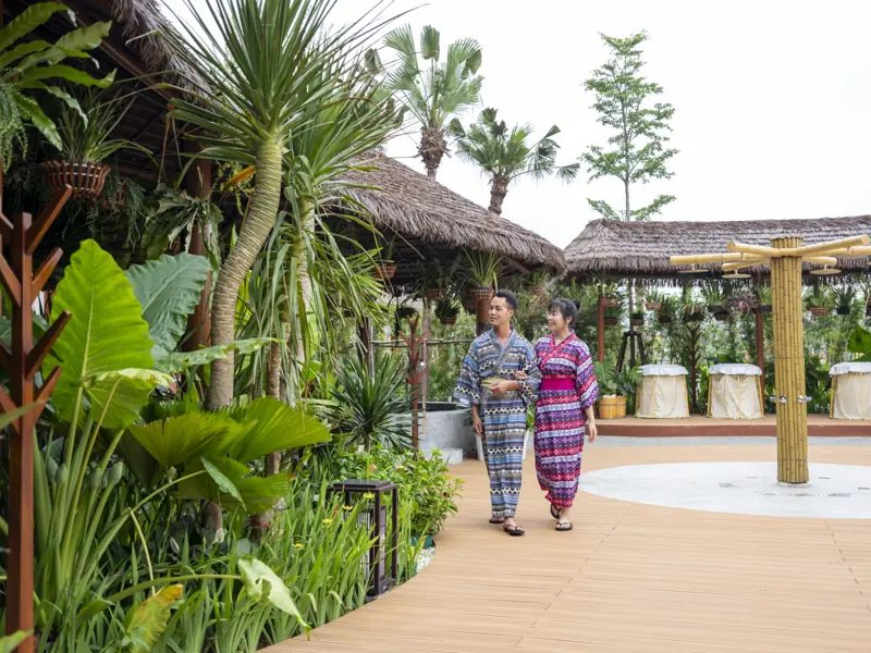 Hai khách mặc áo choàng thổ cẩm đi trên lối gỗ giữa vườn cây nhiệt đới của khu onsen ngoài trời